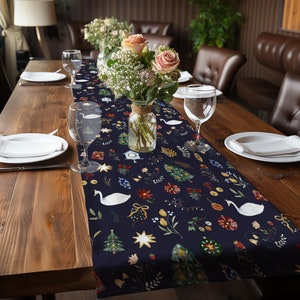 May include: A navy blue table runner with a festive Christmas design featuring white swans, Christmas trees, stars, and other holiday elements. The runner is laid on a wooden table with white plates and glasses.