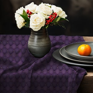 May include: A white bouquet of roses in a gray ceramic vase sits on a purple quilted tablecloth with a single orange on a stack of gray plates.