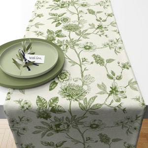 May include: A floral table runner with a cream background and green botanical illustrations. Two green plates with a sprig of greenery and a card that reads "With love," are on the runner. The runner is made of fabric.