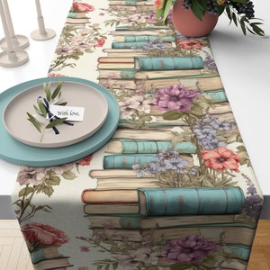 May include: A beige table runner with a floral and book design. The runner features stacks of vintage books with blue and brown spines, and a variety of flowers in shades of pink, purple, and red. The text "With love" is written on a small white card.