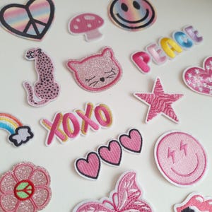 May include: A collection of iron-on patches featuring various designs, including a rainbow heart with a peace sign, a pink mushroom, a pink cat, a pink leopard, a pink star, a pink flower, a pink butterfly, a pink smiley face, a pink heart, and a pink XOXO. The patches are all on a white background.