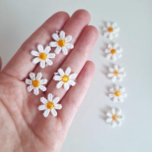 May include: A collection of white embroidered daisy flower patches with yellow centres. The patches are arranged in a line on a white surface.