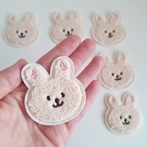 May include: Six white fluffy bunny shaped patches with pink ears and embroidered brown eyes and noses. The patches are approximately 5 cm in diameter.