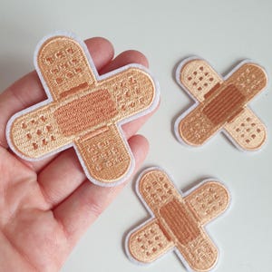 May include: Three embroidered iron-on patches shaped like bandages in a light brown colour.