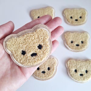 May include: A collection of teddy bear patches. The patches are a light brown colour with a textured, fuzzy appearance. Each bear has black eyes and a black nose. The patches are outlined with a light brown border, on a white background.