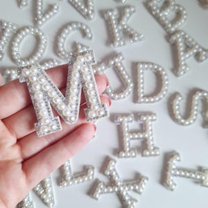 Könnte beinhalten: Ein weißer Buchstabe M aus Perlen und Strasssteinen. Der Buchstabe wird in einer Hand vor einem weißen Hintergrund gehalten, auf dem andere Perlen- und Strassbuchstaben verstreut sind.