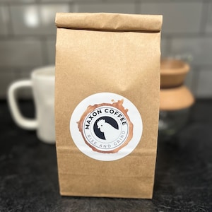 May include: A brown paper bag with a white circle sticker that says "Maxon Coffee Rise and Grind" with a black rooster illustration inside the circle.