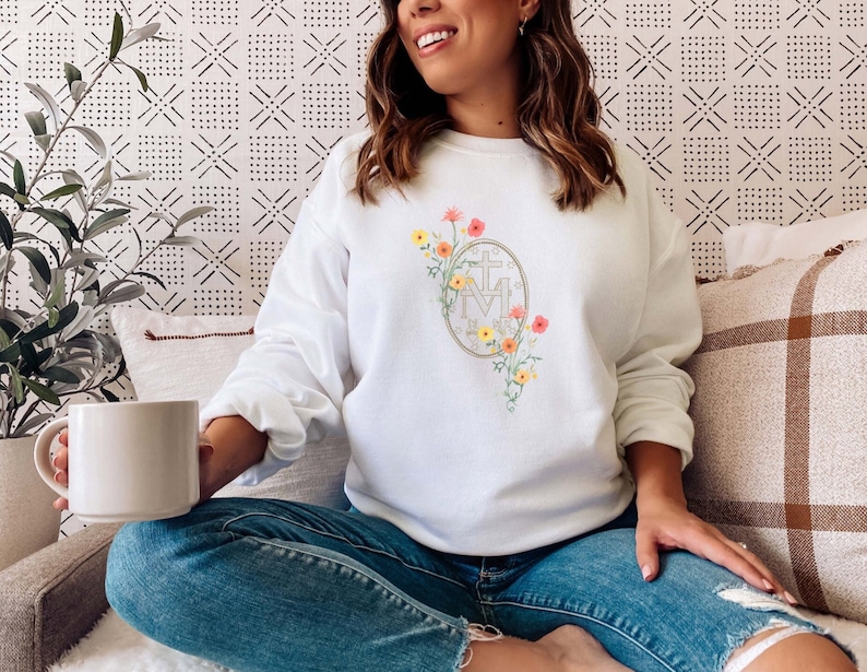 May include: White sweatshirt with a floral design featuring a gold Miraculous Medal. The medal has the letters "M" and "A" with a cross above them. The flowers are yellow, orange, and pink.