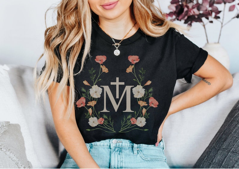 Pu&ograve; includere: Maglietta nera con un design floreale che presenta una croce e la lettera "M" al centro.
