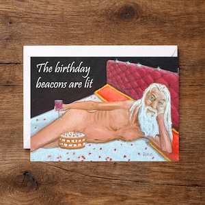 May include: A greeting card featuring an illustration of a bearded figure reclining on a red tufted bed, holding a wine glass. A birthday cake with lit candles sits nearby. The text reads, The birthday beacons are lit, in white script.