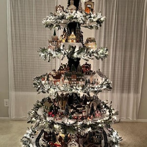May include: A Christmas tree decorated with a miniature village scene. The tree is lit with white lights and has a snowy white base. The village scene includes houses, a church, a train track, and a variety of other buildings.