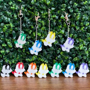 May include: A set of 10 colorful dog keychains. Each keychain features a white dog with a different colored stripe. The keychains are arranged in a rainbow pattern on a wooden surface.