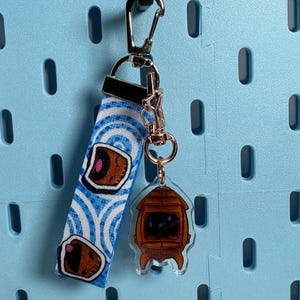 May include: A fabric keychain strap featuring a blue and white wave pattern with cartoon character faces, attached to a silver-tone metal lobster clasp and a small, clear acrylic charm shaped like a brown, rounded, retro-style television set.