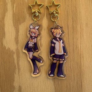 Puede incluir: Dos llaveros acrílicos estilo anime. Cada llavero presenta un personaje con cabello rubio, vistiendo un uniforme blanco y azul marino. Los llaveros están sujetos a cierres dorados en forma de estrella. El fondo es una superficie de madera marrón claro.