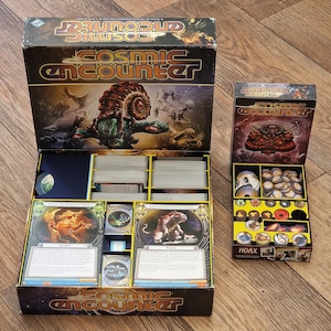 May include: Open board game box titled "Cosmic Encounter" with cards, game pieces, and a smaller box labeled "Cosmic Encounter: Cosmic Eons". The main box features a large alien illustration. The game is set on a wooden surface.