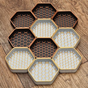May include: Nine hexagonal coasters arranged in a honeycomb pattern. The coasters have a geometric design with a honeycomb pattern in black, copper, and grey. They are displayed on a wooden surface, suitable for drinks.
