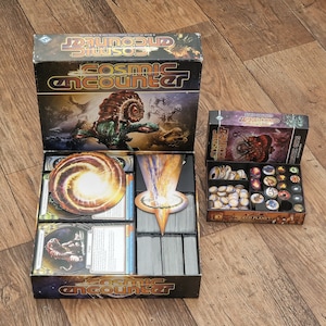 May include: An open box with a Cosmic Encounter board game insert. The insert has compartments for cards, tokens, and other game components. The box lid features the game's title and artwork of a space creature.