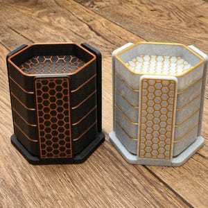 May include: Two hexagonal coasters. One is black with orange accents and a honeycomb pattern. The other is grey with gold accents and a honeycomb pattern. Both have a geometric design and are on a wooden surface.