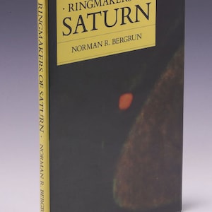 Puede incluir: Un libro titulado "Ringmakers of Saturn" de Norman R. Bergrun. La portada presenta una imagen oscura y abstracta con un pequeño círculo naranja brillante en el centro.