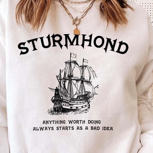 Puede incluir: Sudadera blanca con una ilustración en blanco y negro de un barco y el texto "STURMHOND" sobre el barco. Debajo del barco está el texto "ANYTHING WORTH DOING ALWAYS STARTS AS A BAD IDEA."