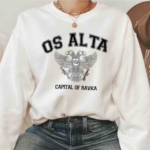 Puede incluir: Sudadera blanca con el texto "OS ALTA" en letras negras sobre un detallado emblema de águila bicéfala. Debajo del emblema, el texto dice "CAPITAL OF RAVKA". La sudadera es de estilo casual.