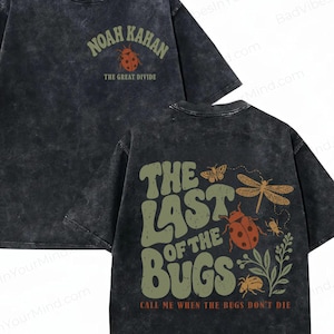 May include: A charcoal grey acid-wash t-shirt featuring a front graphic with the text Noah Kahan and The Great Divide above a ladybug. The back displays the text The Last Of The Bugs and Call Me When The Bugs Don't Die with insect illustrations.