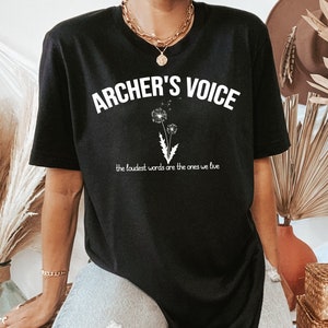 May include: Black t-shirt with the text "ARCHER'S VOICE" in white, above a graphic of two dandelions. Below the text is the phrase "the loudest words are the ones we live". The shirt is a casual style.