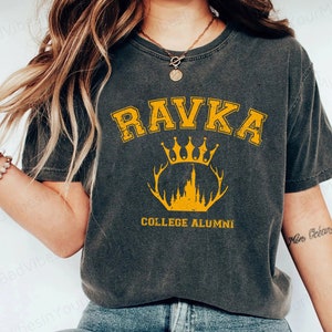 Puede incluir: Una camiseta gris oscura con un diseño gráfico amarillo que dice "RAVKA COLLEGE ALUMNI". El gráfico presenta una corona sobre un horizonte de ciudad estilizado con cuernos a cada lado.
