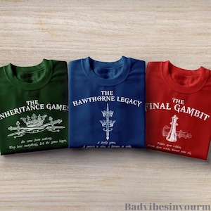 May include: Three t-shirts with different designs. The green t-shirt says "The Inheritance Games" with a crown and swords. The blue t-shirt says "The Hawthorne Legacy" with a crown and sword. The red t-shirt says "The Final Gambit" with a chess piece and the text "Riddle upon riddle, secret upon secret, winner take all."