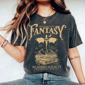 Op de afbeelding: Een donkergrijs T-shirt met een gouden grafisch ontwerp met een draak die over een stapel boeken vliegt. De tekst "Proud Member of the Fantasy Readers Society Dragons, Wizards and Epic Tales" is op het T-shirt gedrukt.