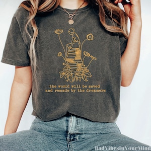 May include: A charcoal gray t-shirt with a yellow graphic of a person sitting on a stack of books with flowers growing from the base of the books. The text "the world will be saved and remade by the dreamers" is printed in yellow below the graphic.