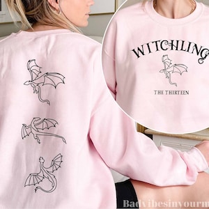 Op de afbeelding: Een roze sweatshirt met een zwarte draak afbeelding en de tekst "Witchling The Thirteen" op de voorkant.