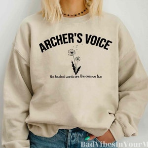 May include: Beige sweatshirt with the text "ARCHER'S VOICE" in black, arched across the top. Below the text is a black and white illustration of a dandelion. The phrase "the loudest words are the ones we live" is printed below the dandelion.