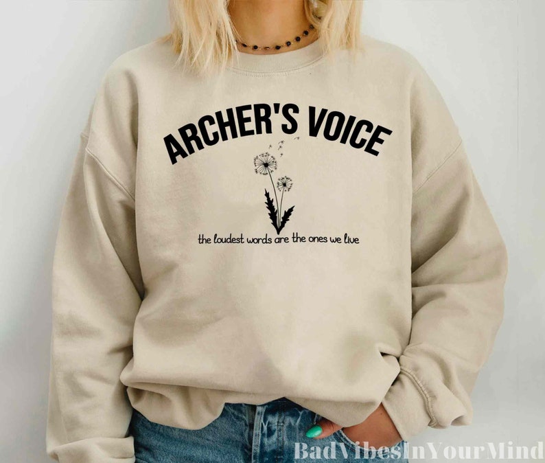 May include: Beige sweatshirt with the text "ARCHER'S VOICE" in black, arched across the top. Below the text is a black and white illustration of a dandelion. The phrase "the loudest words are the ones we live" is printed below the dandelion.