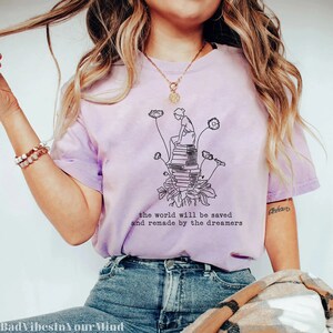 May include: A purple tie-dye t-shirt with a black line drawing of a person sitting on a stack of books with dandelion flowers growing from the base of the stack. The text "the world will be saved and remade by the dreamers" is printed below the drawing.