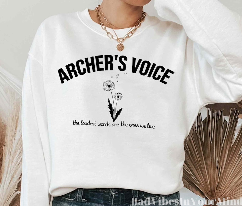 May include: White sweatshirt with black text that reads "ARCHER'S VOICE" and "the loudest words are the ones we live". The text is in a curved arc with a dandelion graphic in the center.