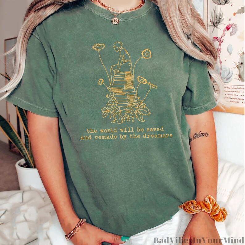May include: A green t-shirt with a yellow graphic of a person sitting on a stack of books with flowers growing from the base of the stack. The text "the world will be saved and remade by the dreamers" is printed below the graphic.