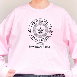 May include: Pink sweatshirt featuring a black graphic of a Camp Half-Blood cabin symbol. The symbol incorporates a Greek key design, an eye, a crescent moon, and a pegasus. The text "Camp Half-Blood Cabin Fifteen Hypnos Long Island Sound" is printed beneath the symbol.