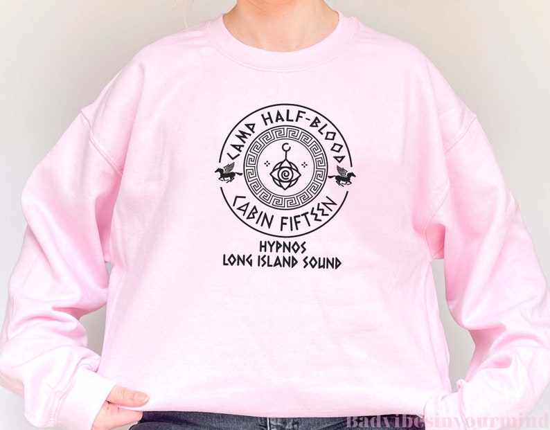 May include: Pink sweatshirt featuring a black graphic of a Camp Half-Blood cabin symbol. The symbol incorporates a Greek key design, an eye, a crescent moon, and a pegasus. The text "Camp Half-Blood Cabin Fifteen Hypnos Long Island Sound" is printed beneath the symbol.