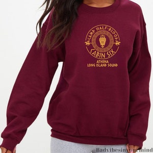 May include: Burgundy sweatshirt with a gold graphic of a Camp Half-Blood logo. The logo features an owl, a Greek key design, and the text "Camp Half-Blood Cabin Six Athena Long Island Sound".