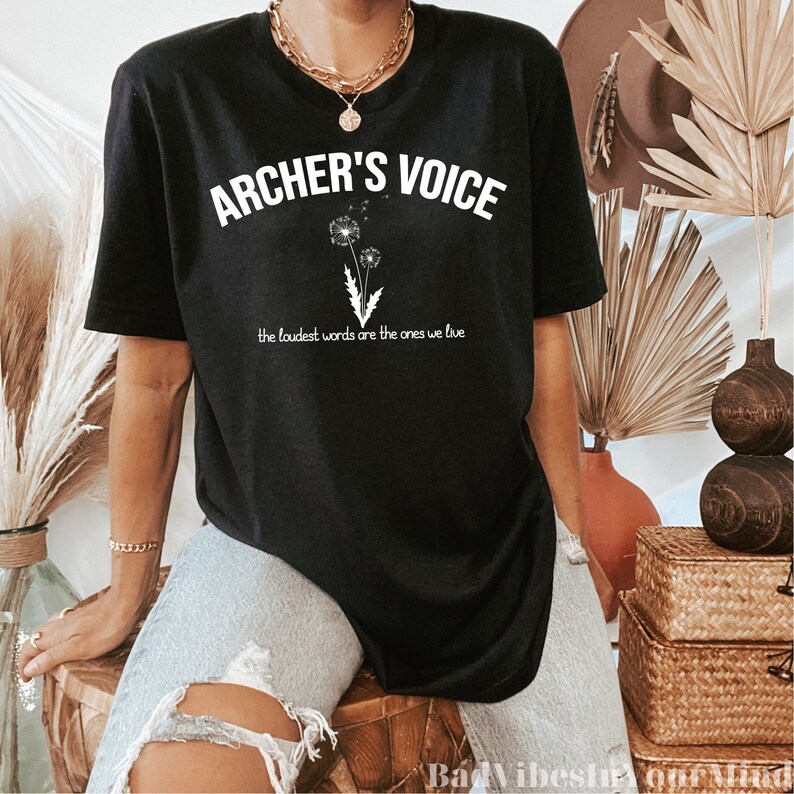May include: Black t-shirt with the text "ARCHER'S VOICE" in white, above a graphic of two dandelions. Below the text is the phrase "the loudest words are the ones we live". The shirt is a casual style.