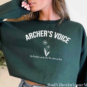 May include: A dark green sweatshirt with the text "ARCHER'S VOICE" in white, along with a dandelion graphic. Below the text is the phrase "the loudest words are the ones we live".