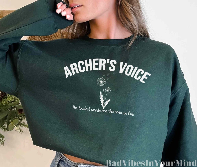 May include: A dark green sweatshirt with the text "ARCHER'S VOICE" in white, along with a dandelion graphic. Below the text is the phrase "the loudest words are the ones we live".