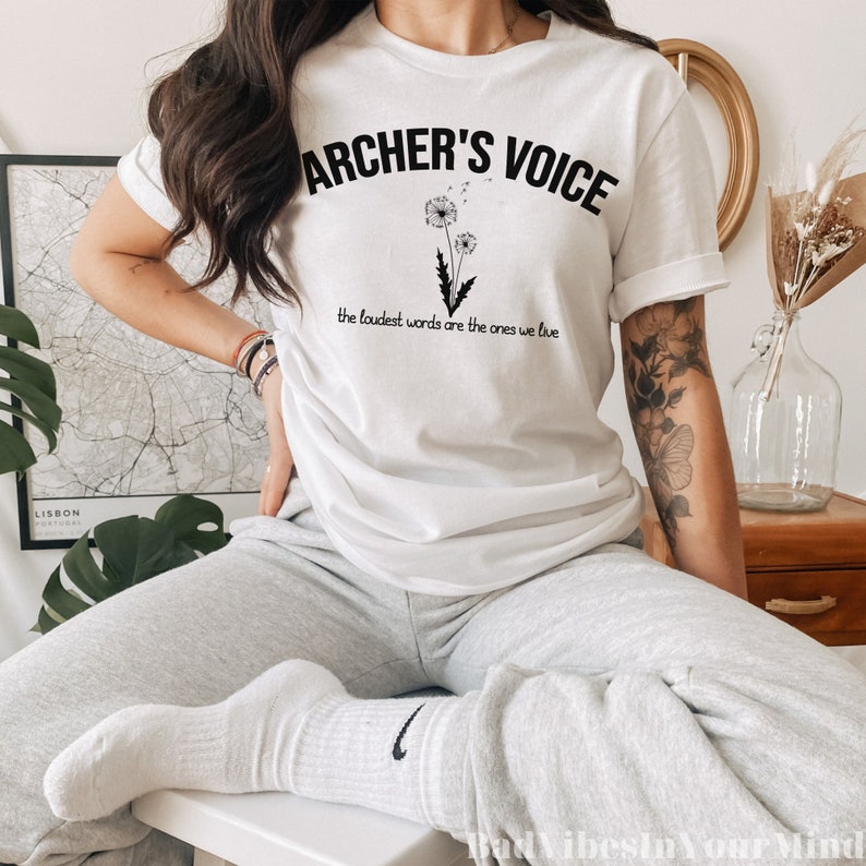 May include: White t-shirt with the text "ARCHER'S VOICE" above a dandelion graphic. Below the graphic, the text reads "the loudest words are the ones we live". The shirt is paired with gray sweatpants and white socks.