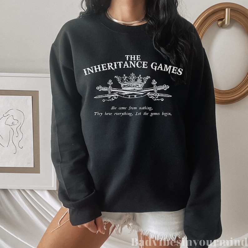 May include: Black sweatshirt with white text that reads "THE INHERITANCE GAMES" with a crown and swords graphic. The text below the graphic reads "She came from nothing. They have everything. Let the games begin."
