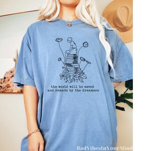 May include: A light blue t-shirt with a black line drawing of a person sitting on a stack of books with flowers growing around them. The text "the world will be saved and remade by the dreamers" is printed below the drawing. The words "Bad Vibes In Your Mind" are printed at the bottom of the shirt.