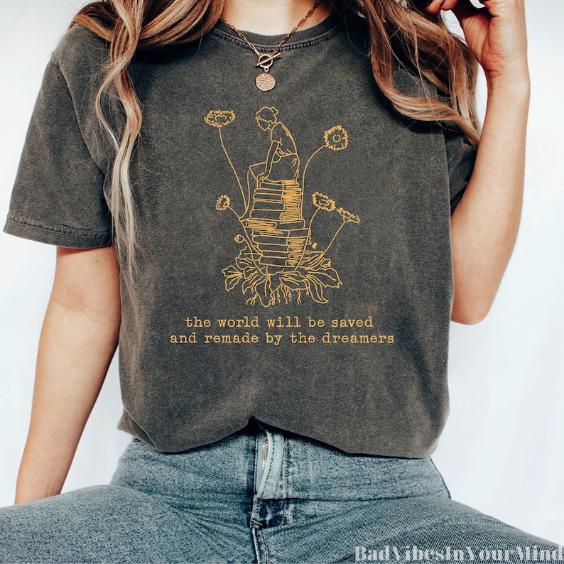 May include: A charcoal gray t-shirt with a yellow graphic of a person sitting on a stack of books with flowers growing from the base of the books. The text "the world will be saved and remade by the dreamers" is printed in yellow below the graphic.