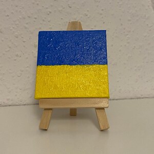 May include: A small canvas painting of the Ukrainian flag on a wooden easel. The flag is blue on top and yellow on the bottom.