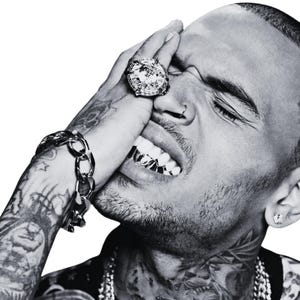 May include: A black and white portrait of a person with extensive arm and neck tattoos, wearing a large diamond ring on their finger, a thick metallic chain bracelet, a nose ring, metallic teeth grills, and layered silver chains around their neck.