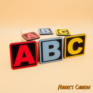May include: Five wooden blocks with black borders and colorful letters spelling out "ABC" in a playful design. The blocks are arranged in a row on a wooden surface.